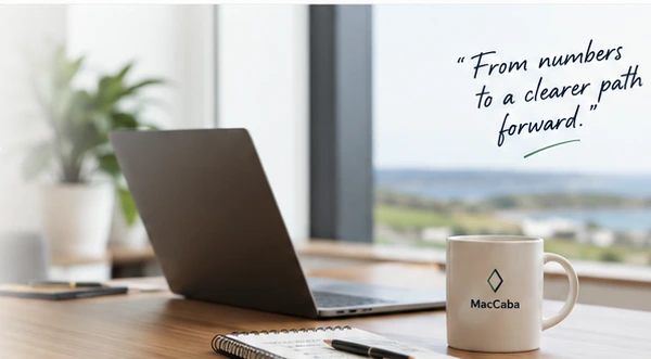 A desk with a laptop, notebook and MacCaba mug near a bright window.